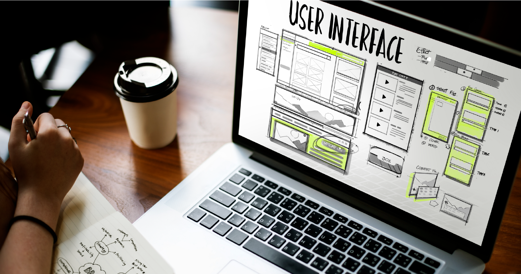 Designer sketching user interface wireframes on a laptop with UI layout elements, next to a notebook and coffee cup on a wooden desk.